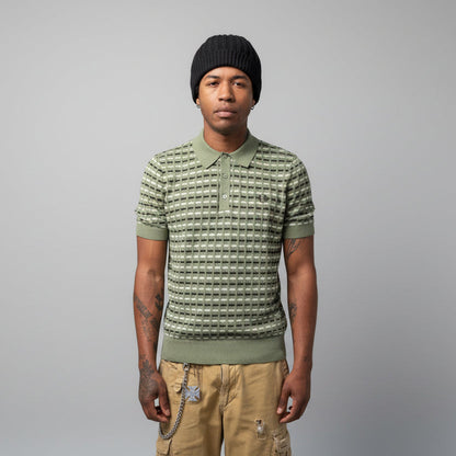 A person wearing a Fred Perry boucle ribbed knitted shirt in olive mint color, paired with a beanie and shorts, standing in front of a wall with graffiti.