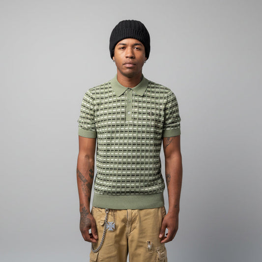 A person wearing a Fred Perry boucle ribbed knitted shirt in olive mint color, paired with a beanie and shorts, standing in front of a wall with graffiti.
