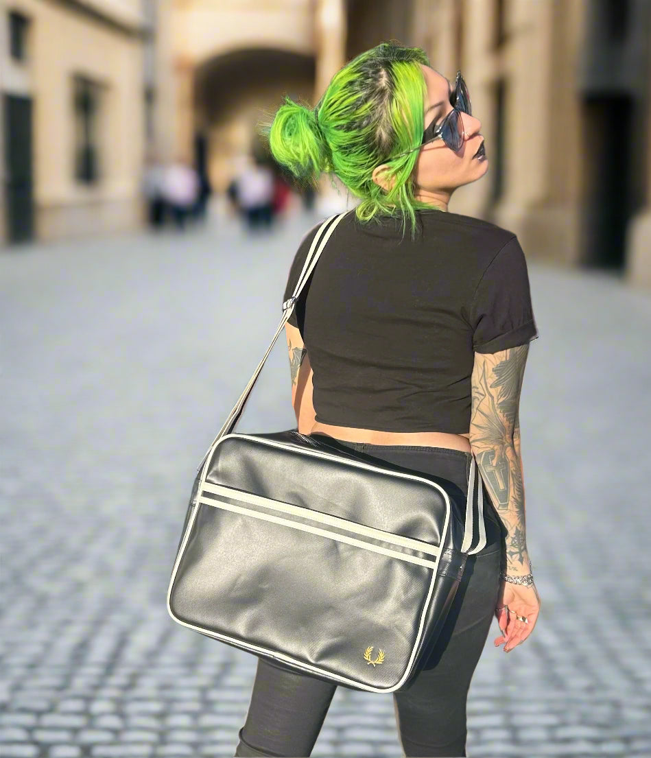 A person with green hair and sunglasses is seen from behind, wearing a black crop top and carrying a silver Fred Perry classic shoulder bag.
