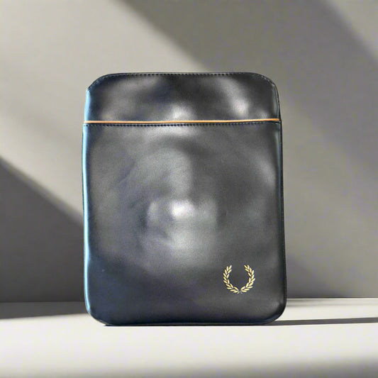 A black Fred Perry leather iPad cover displayed in a shadowed setting, highlighting its sleek design and the brand's logo on the bottom corner.