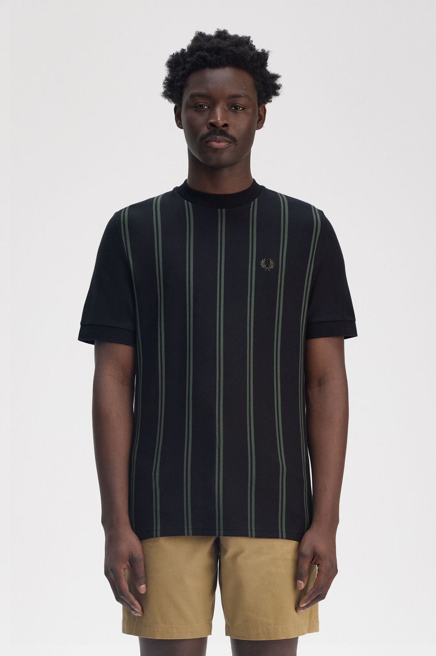 A male model wearing a black Fred Perry striped t-shirt with a V-neck collar and short sleeves, paired with brown trousers.