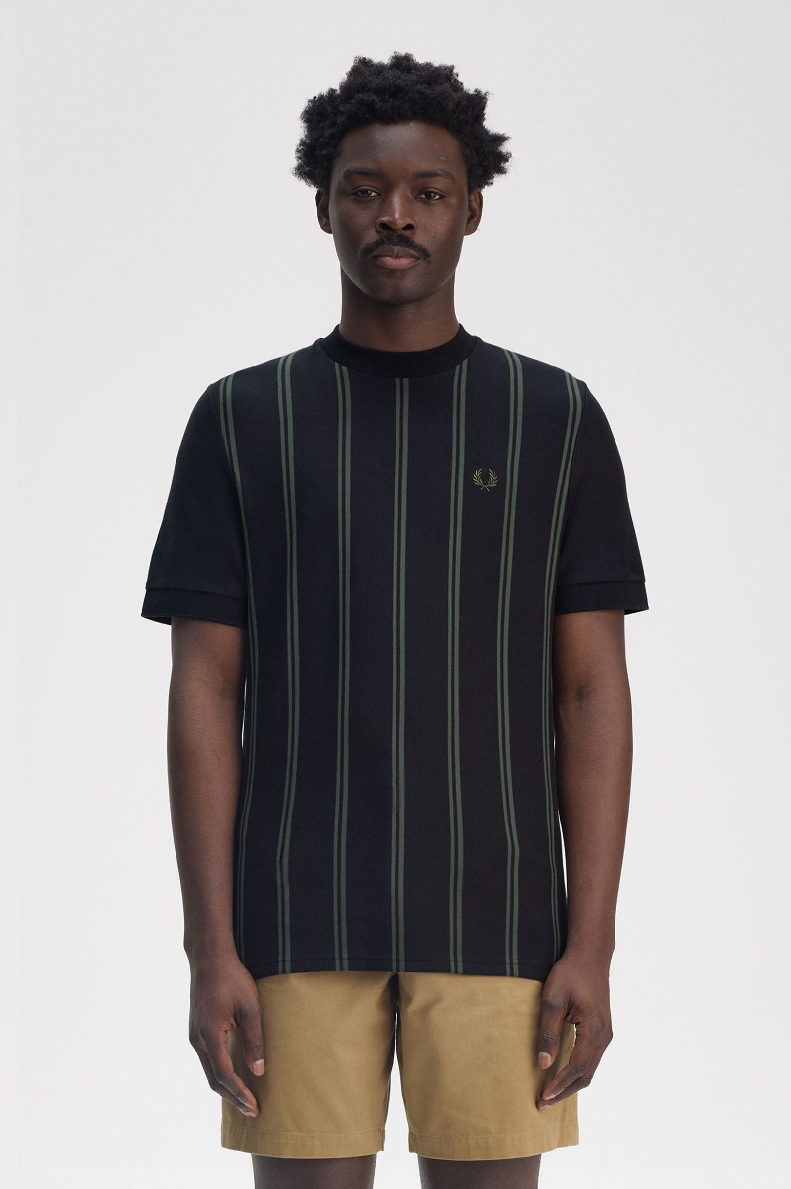 A male model wearing a black Fred Perry striped t-shirt with a V-neck collar and short sleeves, paired with brown trousers.