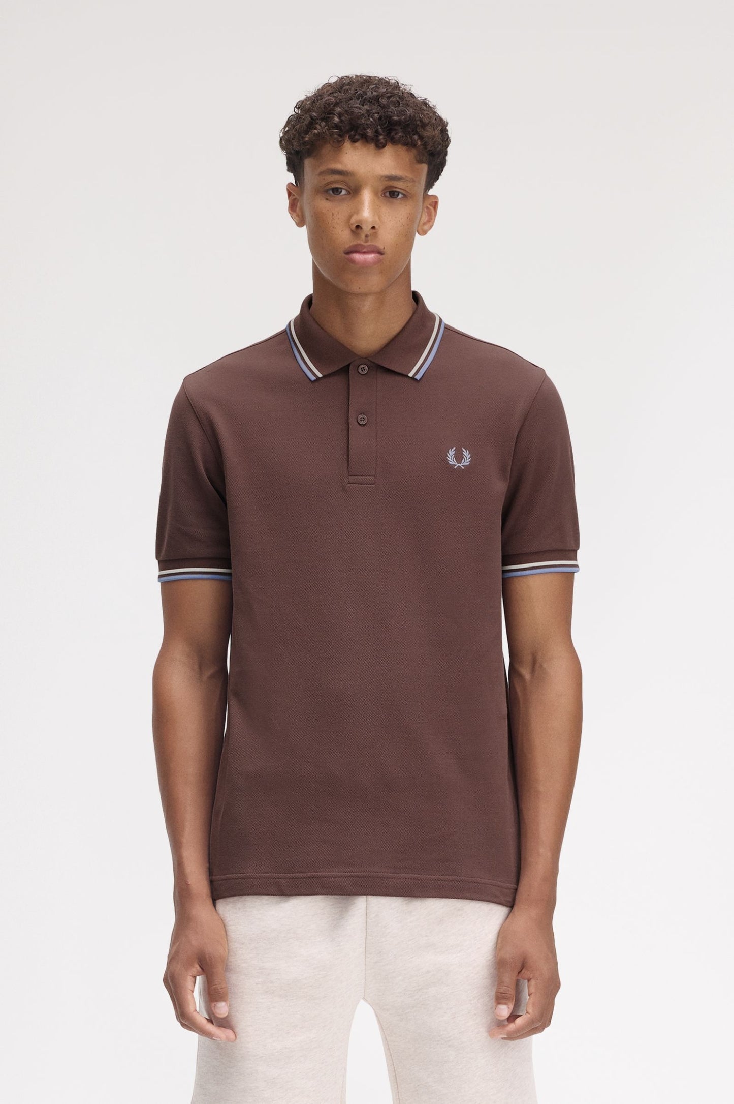A man wearing a brown Fred Perry twin tipped polo shirt with white trim on the collar and sleeves, paired with white pants.