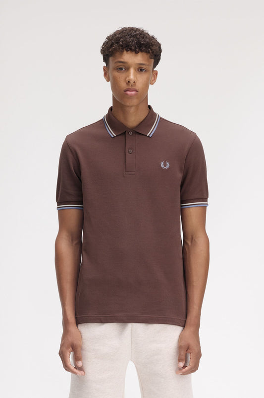 A man wearing a brown Fred Perry twin tipped polo shirt with white trim on the collar and sleeves, paired with white pants.