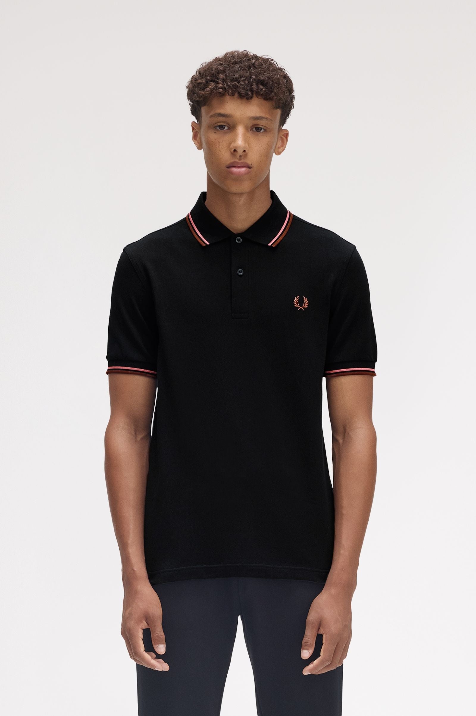 A man wearing a brown Fred Perry twin tipped polo shirt with white trim on the collar and sleeves, paired with white pants.