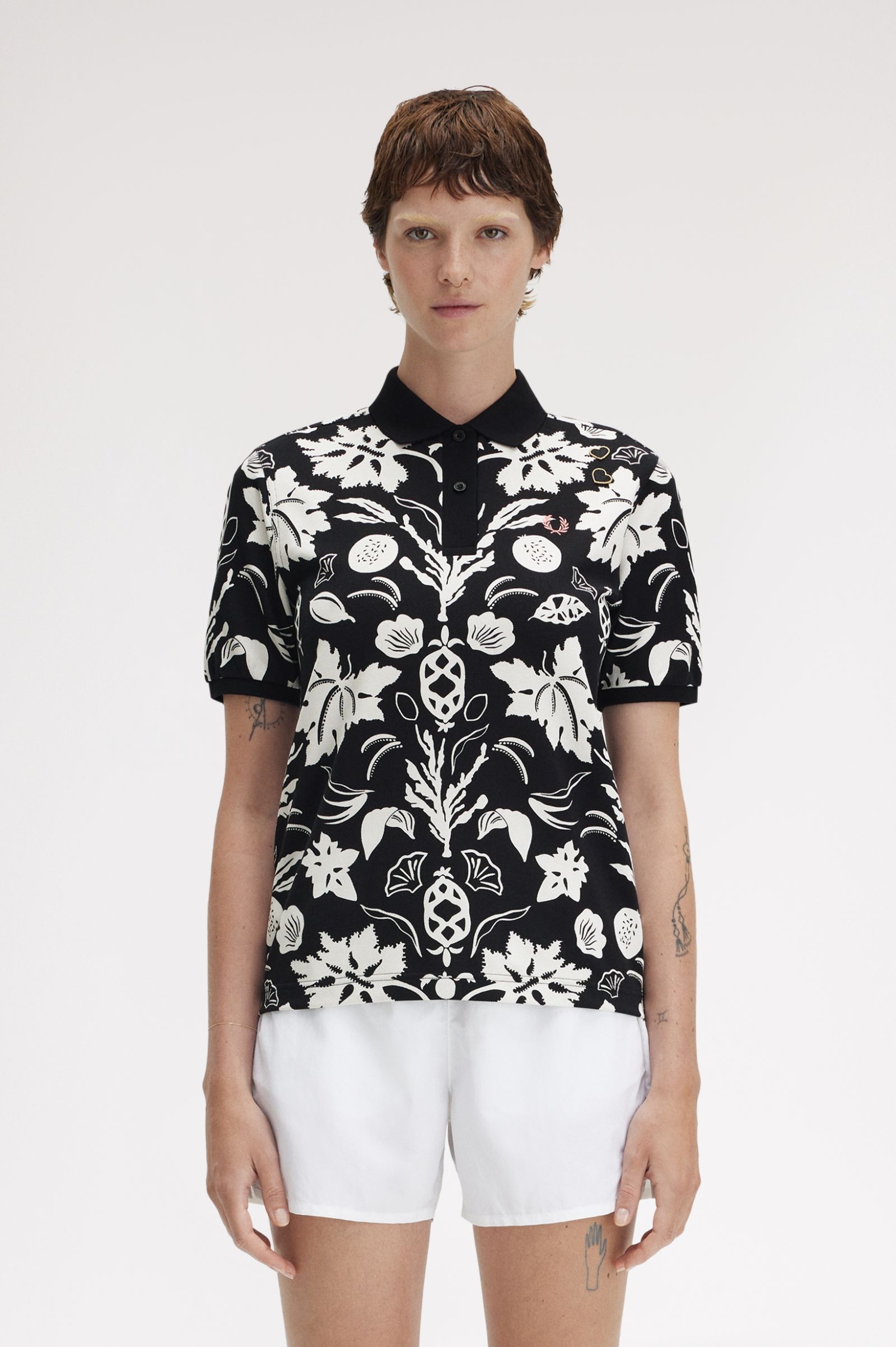 A person wearing a black and white floral printed Fred Perry shirt with short sleeves, paired with white shorts.