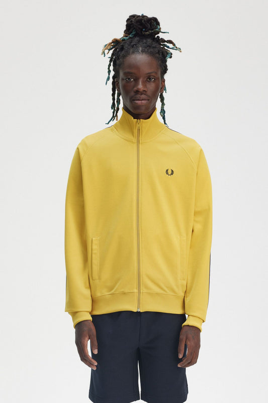 A person wearing a yellow Fred Perry track jacket with black tape on the sleeve, paired with dark shorts.