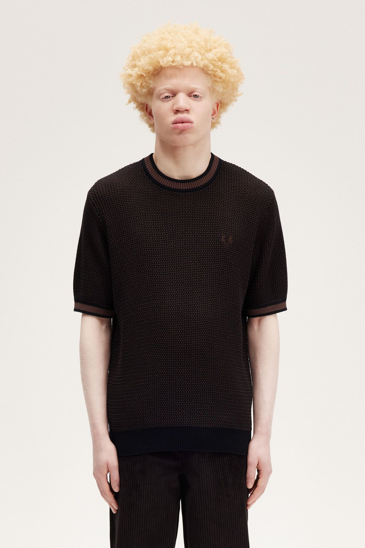 A male model wearing a black textured knitted t-shirt with a crew neck and two-tone construction. The shirt is displayed on a white background.