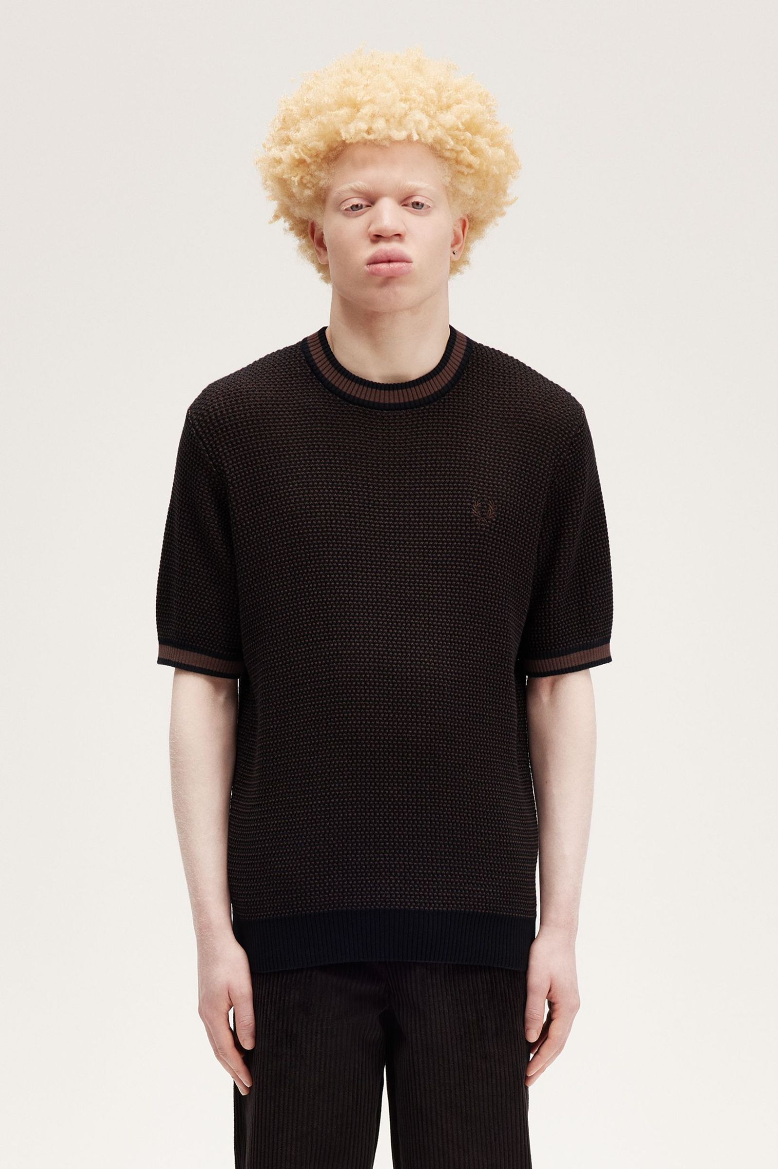 A male model wearing a black textured knitted t-shirt with a crew neck and two-tone construction. The shirt is displayed on a white background.