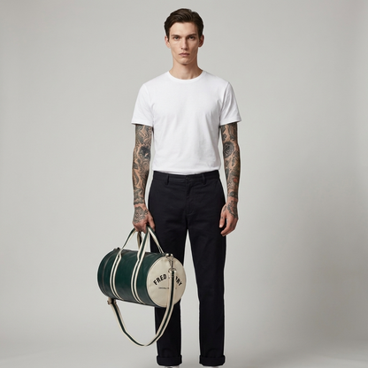 A male model with tattooed arms stands against a light gray studio background, wearing a white crew-neck t-shirt and dark trousers while holding a green and cream cylindrical duffel bag with the brand name 'FREDERICK PECK' printed on it.