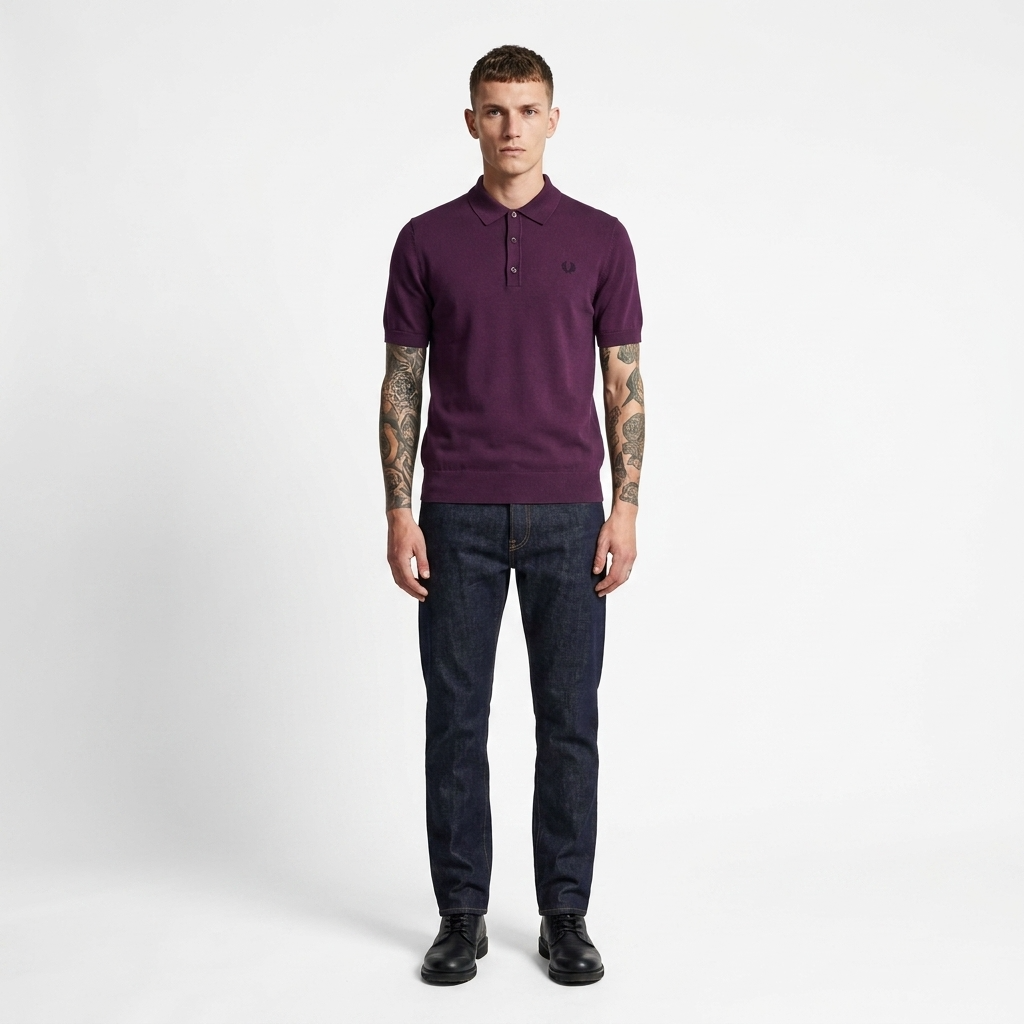 A man wearing a dark purple polo shirt with a small embroidered logo on the chest, paired with dark blue jeans and black shoes, standing against a light gray studio background.