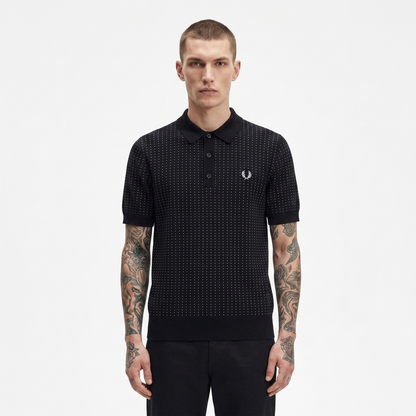 A man modeling a black short-sleeve polo shirt with a subtle micro-dot pattern and the iconic embroidered laurel branch logo on the chest, set against a plain light gray background.