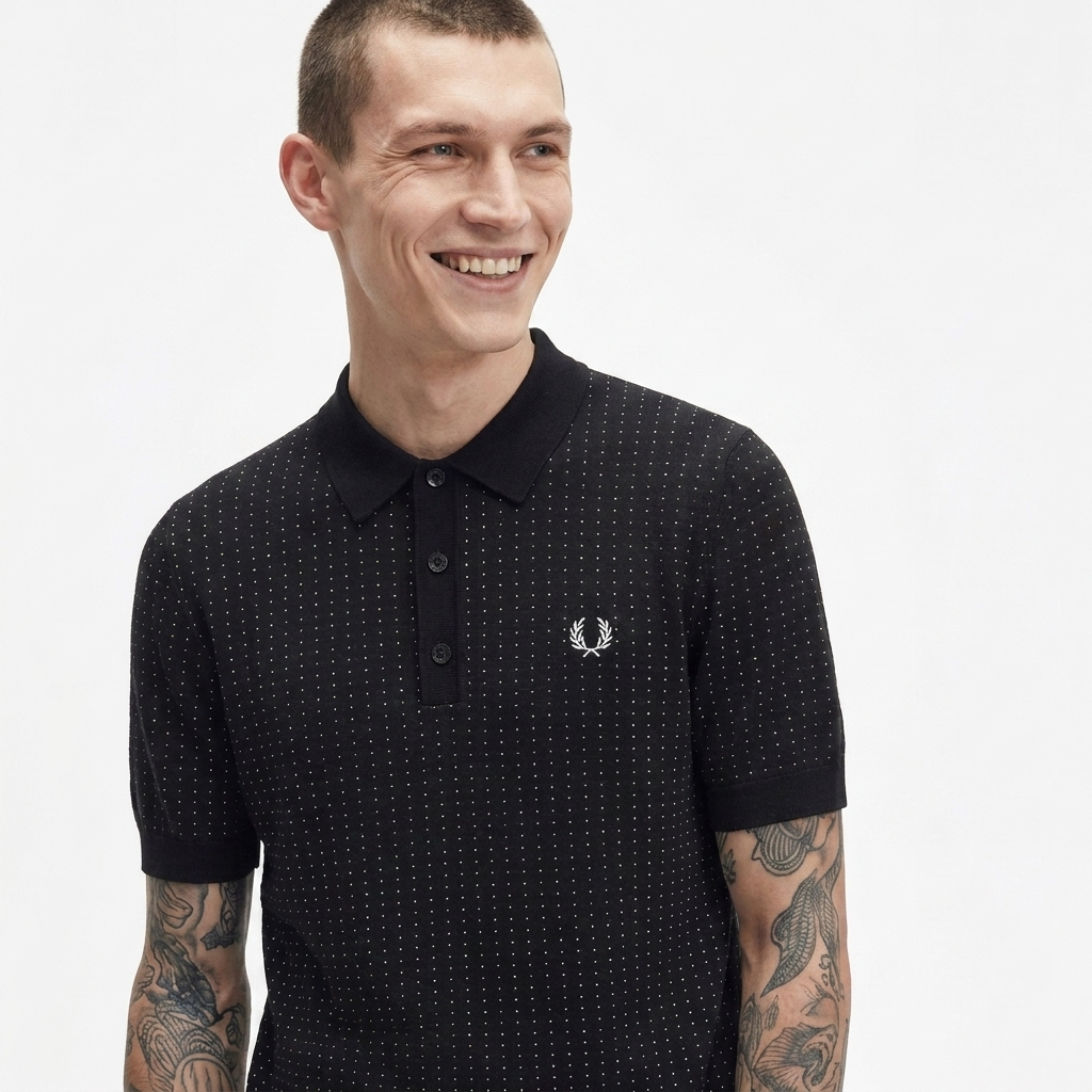 A smiling man wearing a black short-sleeve polo shirt with a subtle white micro-dot pattern and the embroidered white Fred Perry laurel wreath logo on the chest, set against a plain light gray background.