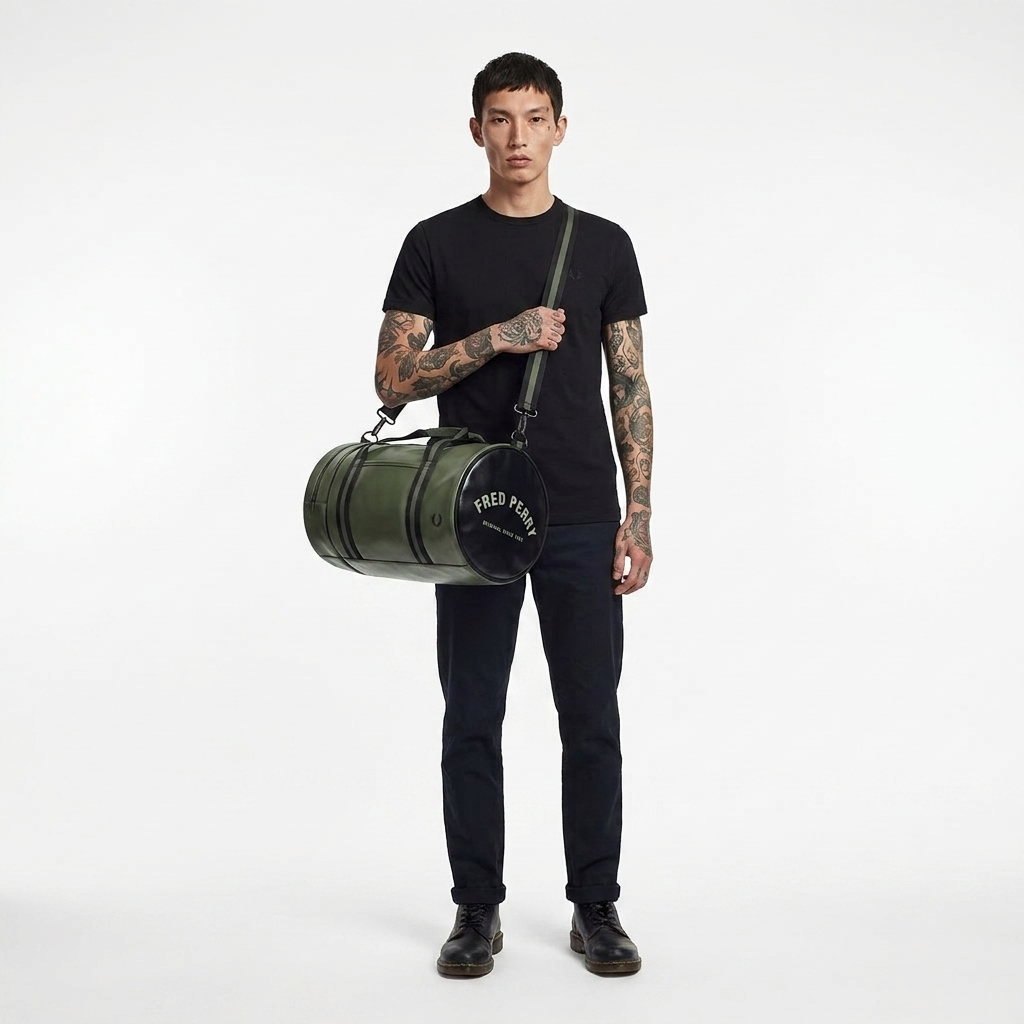 A man with tattoos stands against a white studio background wearing a black t-shirt, dark jeans, and black shoes, while carrying a green cylindrical duffel bag with a black top and white 'FRED PERRY' branding.