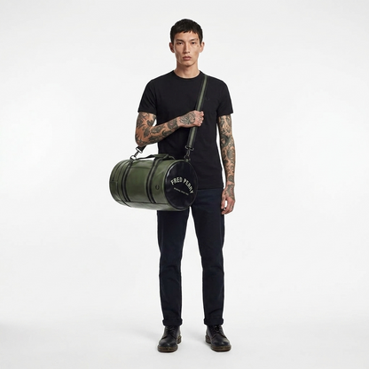 A man with tattoos stands against a white studio background wearing a black t-shirt, dark jeans, and black shoes, while carrying a green cylindrical duffel bag with a black top and white 'FRED PERRY' branding.