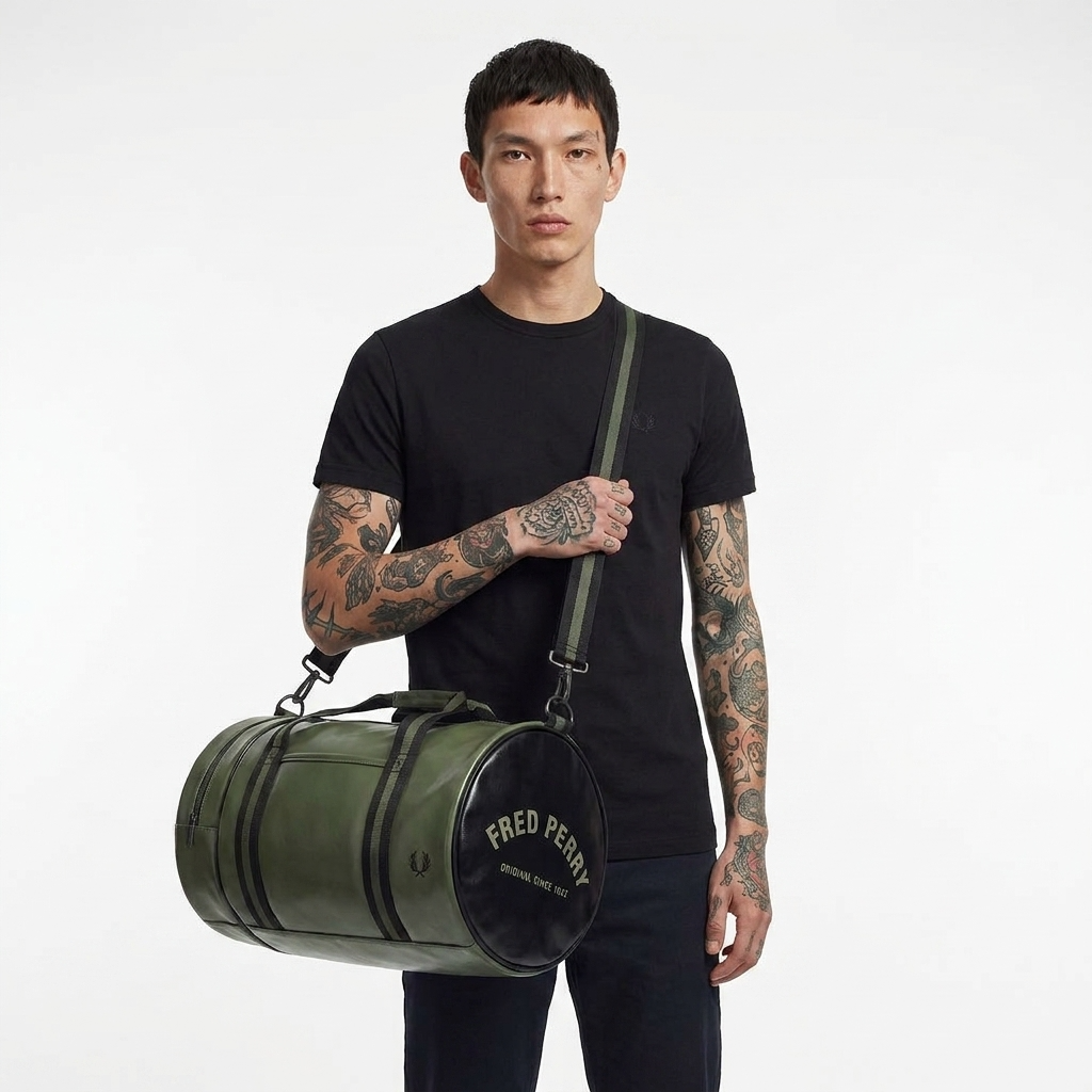 A model with tattooed arms wearing a black t-shirt and dark pants, holding a green cylindrical duffel bag with a black circular end-cap that reads 'FRED PERRY'.