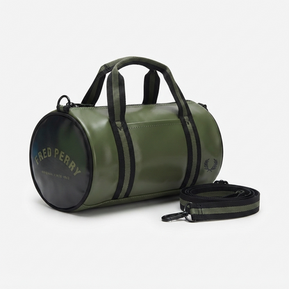 An olive green cylindrical duffel bag with black handles and side stripes, featuring a black circular end cap with gold 'FRED PERRY' branding and a matching black-and-green shoulder strap on a light gray background.