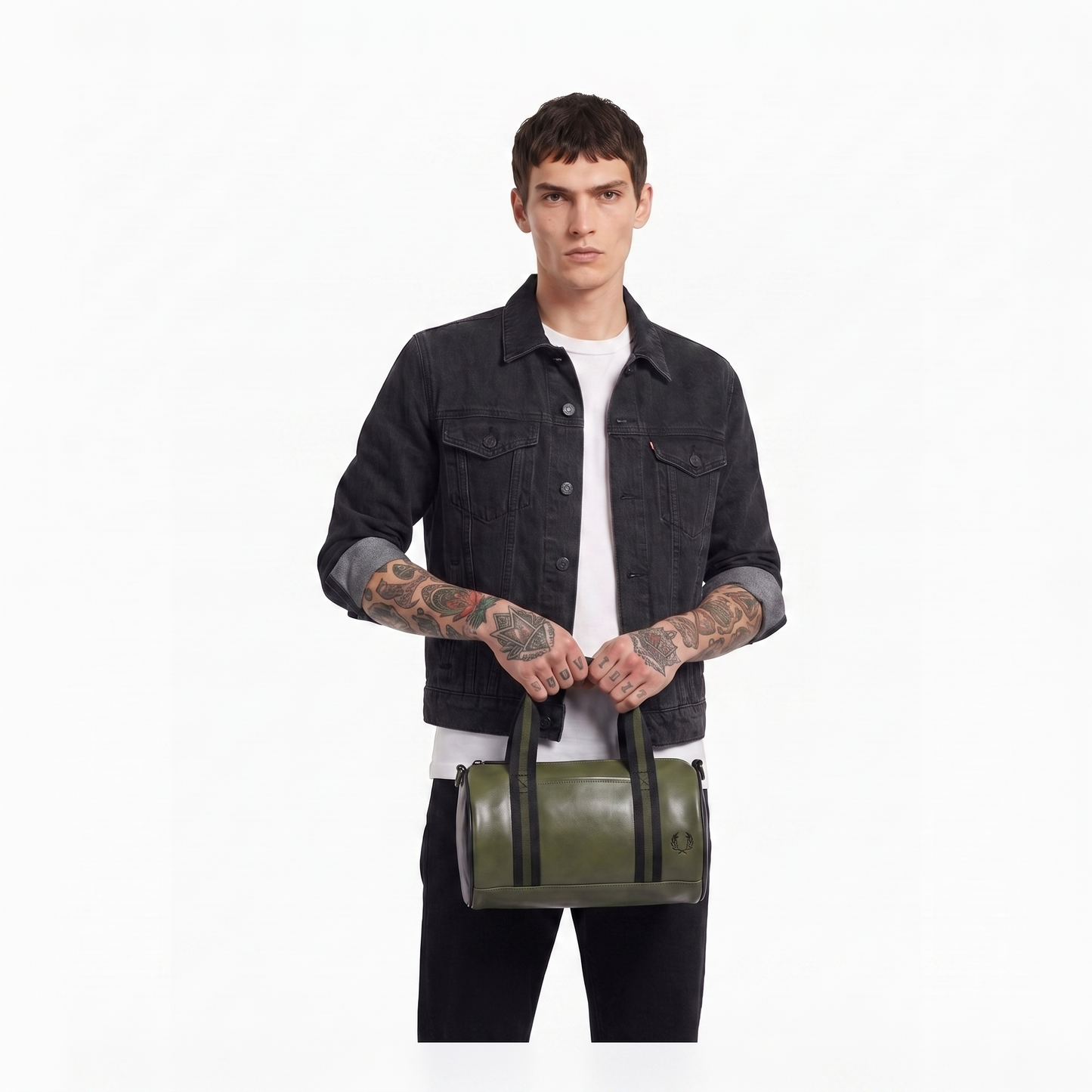 A model wearing a black denim jacket and white t-shirt holds an olive green cylindrical duffel bag with black handles against a plain white background.