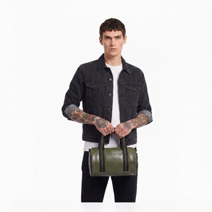 A model wearing a black denim jacket and white t-shirt holds an olive green cylindrical duffel bag with black handles against a plain white background.
