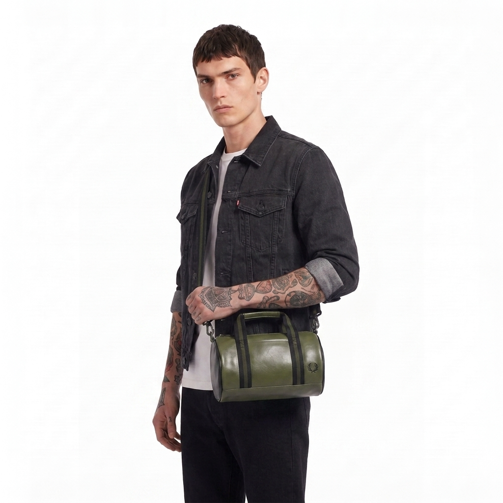 A man wearing a dark denim jacket and black pants carries an olive green leather duffel bag with black straps and handles against a plain white background.