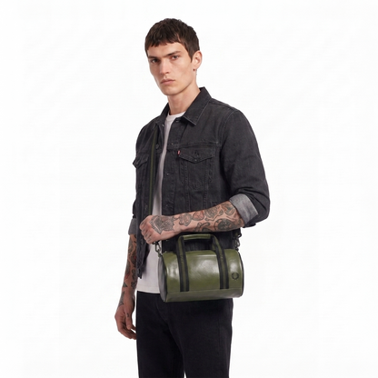 A man wearing a dark denim jacket and black pants carries an olive green leather duffel bag with black straps and handles against a plain white background.