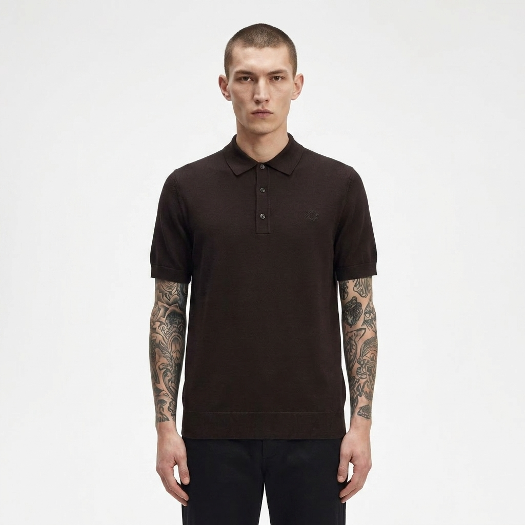 A man with short hair and visible arm tattoos wearing a dark brown short-sleeve polo shirt, standing against a plain light gray studio background.