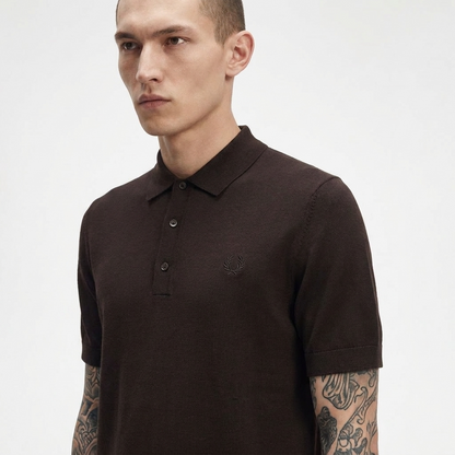 A man wearing a dark brown short-sleeve polo shirt with a small embroidered logo on the chest, photographed against a light gray studio background.