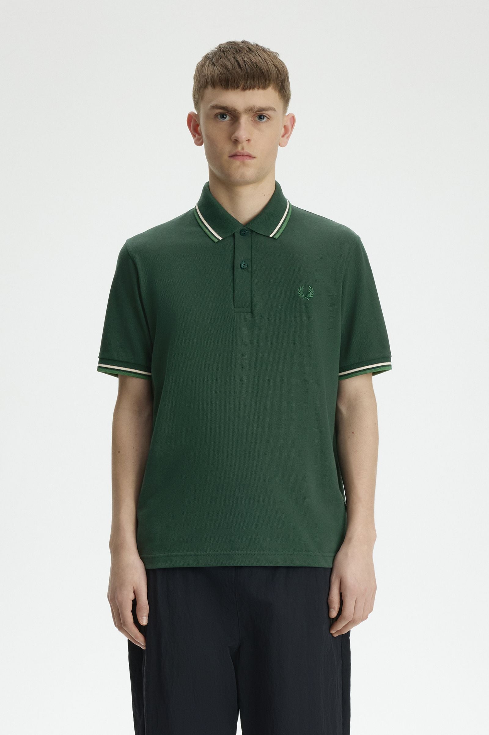 A man wearing a dark green short-sleeve polo shirt with white contrast piping on the collar and sleeve cuffs, paired with dark trousers, photographed against a light gray studio background.