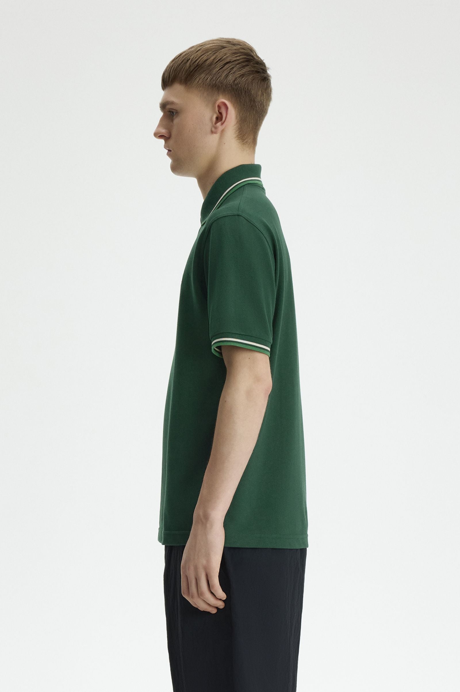 A side-profile view of a young man wearing a dark green short-sleeve polo shirt with thin white stripes on the collar and sleeve cuffs, paired with dark trousers, against a plain light gray background.