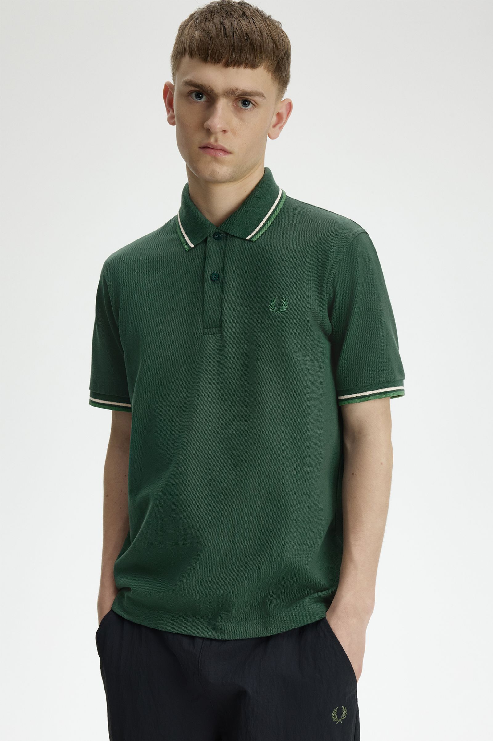 A man wearing a dark green short-sleeve polo shirt with white piping on the collar and sleeve cuffs, paired with dark trousers, photographed against a light gray studio background.