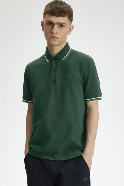 A man wearing a dark green short-sleeve polo shirt with white piping on the collar and sleeve cuffs, paired with dark trousers, photographed against a light gray studio background.