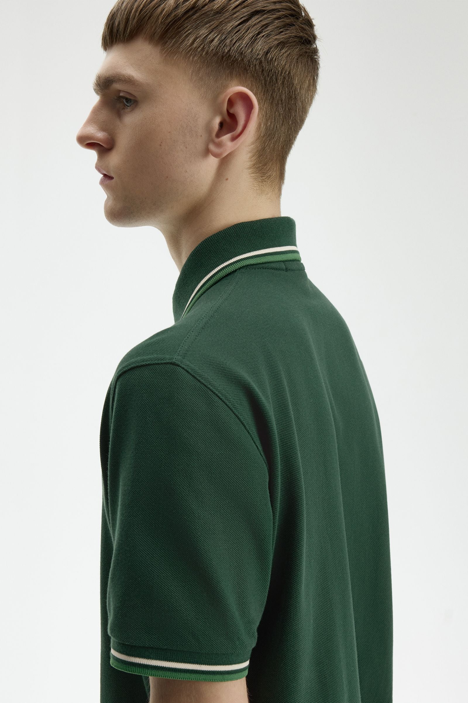 A man shown in profile wearing a dark green short-sleeve polo shirt with white stripe trim on the collar and sleeve cuffs, against a plain light gray studio background.