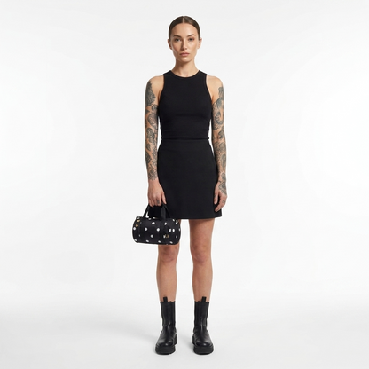 A model with tattoos stands against a light gray studio background wearing a sleeveless black mini dress, black ankle boots, and holding a small black handbag with white polka dots.