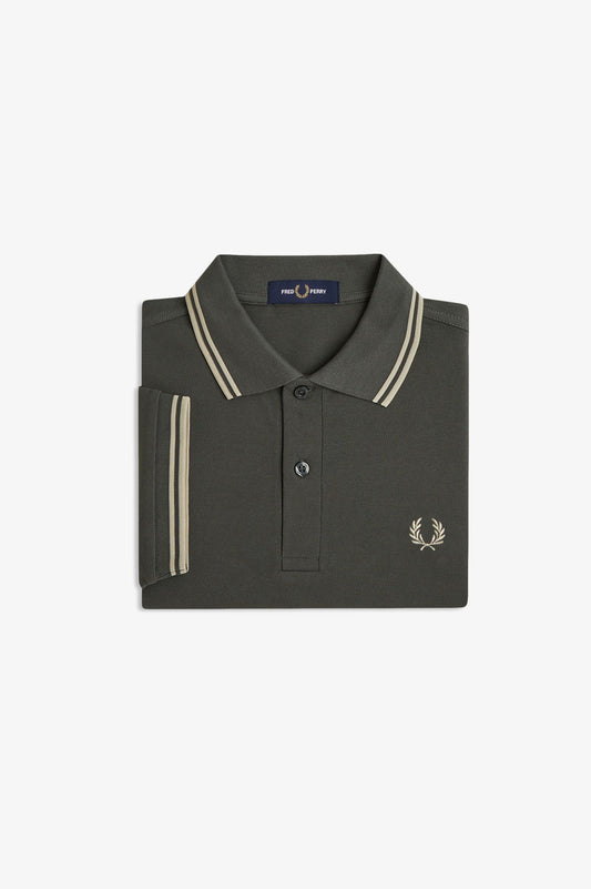 A folded dark olive green polo shirt by Fred Perry, featuring cream-colored double stripes on the collar and sleeve cuffs, and the signature embroidered laurel wreath logo on the chest.