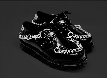 A pair of black leather lace-up sneakers with double chains and spiked detailing, displayed against a dark background.